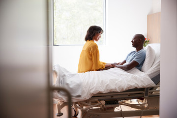 Wife Visiting And Talking With Patient Husband In Hospital Bed