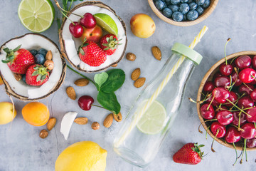 Top view Clear-colored cooling drink in a glass bottle with a straw, a number of broken coconut, strawberries, cherries, apricots, blueberries, lime, lemon, nuts on a dark stone background, save space