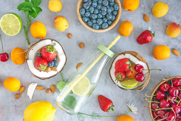 Top view Clear-colored cooling drink in a glass bottle with a straw, a number of broken coconut, strawberries, cherries, apricots, blueberries, lime, lemon, nuts on a dark stone background, save space