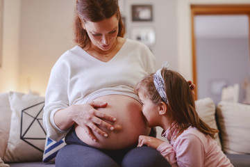 Obraz premium Cute little girl kissing mother's belly. Living room interior.