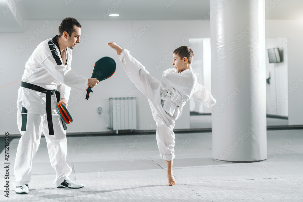 Wall mural Young Caucasian boy in dobok kicking barefoot while trainer holding kick target. Taekwondo training concept.