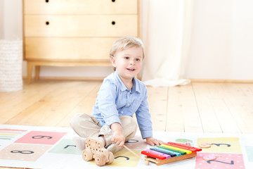 The boy plays xylophone at home. Cute smiling positive boy playing with a toy musical instrument xylophone in the children's white room. Close-up of kid playing on xylophone. Child development concept