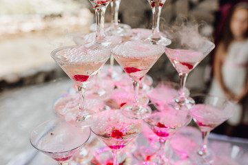 Dry Ice Martini Glass Pyramid with Red Cherry on Wedding Table. Beautiful Delicious Alcohol Crystal Tower with Champagne Outdoor Background. Romantic Celebrations and Party Catering Concept