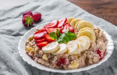 Oatmeal porridge with milk, strawberry, banana in white plate on wooden table background