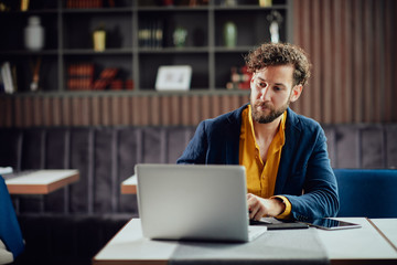 Young serious bearded businessman in smart casual clothes sitting in cafe and typing on laptop.