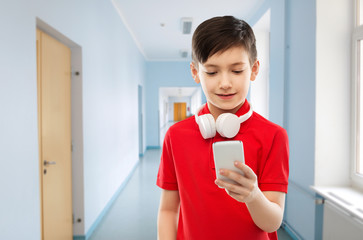education, technology and people concept - smiling little boy in red polo t-shirt with headphones on neck using smartphone over school corridor background