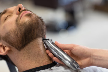 grooming and people concept - man and barber with trimmer or shaver cutting beard at barbershop