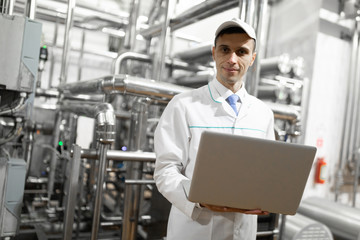Naklejka na ściany i meble Portrait of man in a white robe and a cap standing in production department of dairy factory with laptop