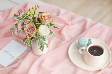 French macaroon blue plate on the pink and coffee cup standing on a wooden table with a pink tablecloth white vase with flowers roses and greens.