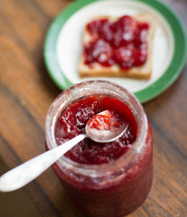 Jar of strawberry jam with the spoon in it, and a slice of bread with strawberry jam served on the kitchen counter