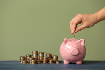 Female hand putting coin into piggy bank on table against color background. Money savings concept