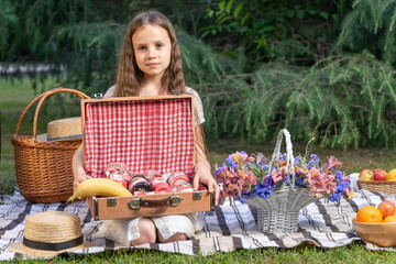 Portrait of a beautiful girl with a basket of flowers on a picnic in the park. The child holds open a chest with jam