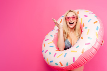 Obraz na płótnie Canvas Image of girl in swimsuit sunglasses with inflatable donut for swimming on empty pink background. Indicates with hand to side