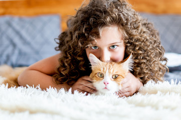 Cute girl with curly hair hugs a red cat on the bed..