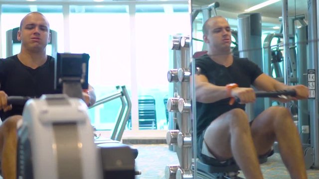 Young athlete makes an exercise on rowing simulator in gym reflecting in mirror