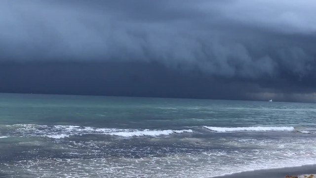 Hurricane with thick ominous clouds is seen over the ocean approaching a beach. There is a boat in the distance on the calm ocean water