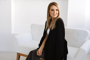 Portrait of happy smiled blond young girl is wearing black suit, sitting on white sofa on white background. Place for text