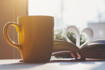 open book with pages shaped as heart and yellow coffee cup on wooden table, love story, education...