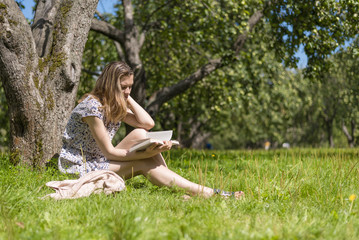 Naklejka na ściany i meble Young attractive happy woman reading a book in a rural apple orchard - the concept of simple village life