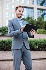 Young, handsome businessman working on a tablet in front of an office building. Low angle view. Work and stay connected anywhere concept