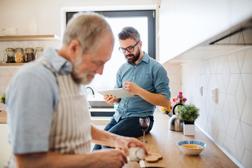 Adult hipster son and senior father indoors in kitchen at home, using tablet.