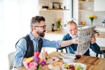 An adult hipster son and senior father indoors at home, eating light lunch.