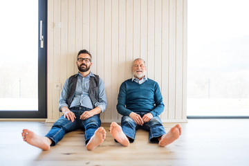 A portrait of adult hipster son and senior father sitting on floor indoors at home.
