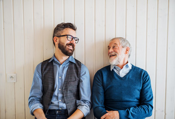 A portrait of adult hipster son and senior father sitting on floor indoors at home.