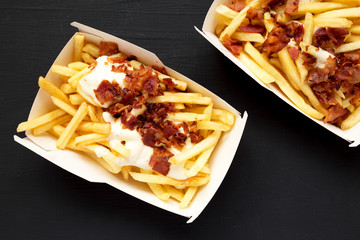 Top view, french fries with cheese sauce and bacon in paper boxes over black background. Flat lay,...