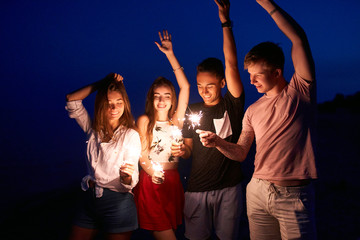 Friends walking, dancing and having fun during night party at the seaside with bengal sparkler lights in their hands. Young teenagers partying on the beach with fireworks. Slow motion steadycam shot.