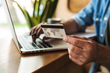 Photo cropped of european man holding credit card and typing on laptop while working in cafe indoors