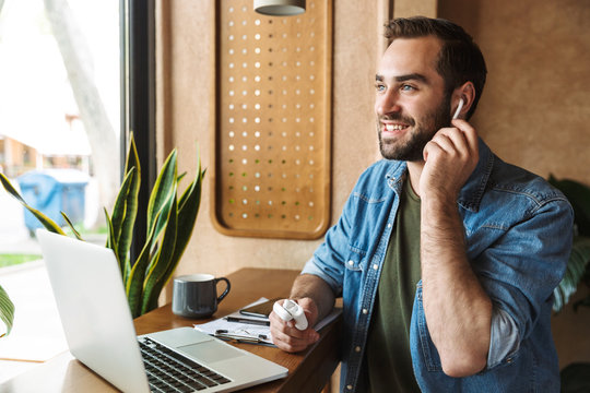Photo of laughing optimistic man using earpods with laptop and clipboard while working in cafe indoors