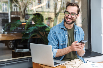 Photo of brunette happy man typing on cellphone with laptop while working in cafe outdoors