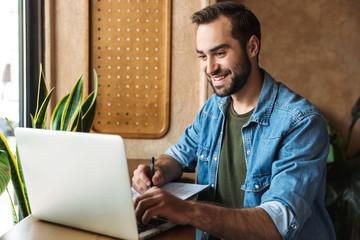 Photo of brunette bearded man writing and typing on laptop while working in cafe indoors