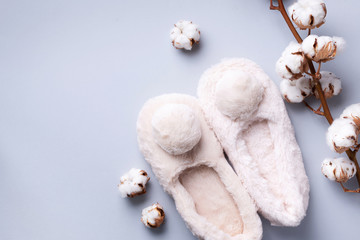 Nude fluffy home slippers with pompon and cotton flower branch on grey background with copy space. Top view. Copy space. Cozy home footwear. Sweet home and spa, body treatment concept