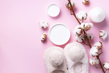 Bathroom accessories, nude fluffy home slippers, cotton flowers, candle, skin care products on pink background with copy space. Top view. Copy space. Spa and body treatment concept.