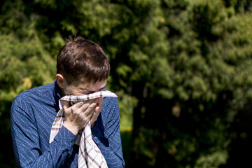 Closeup portrait of a young man with allergies. A man sneezes into a scarf on the background of green trees.