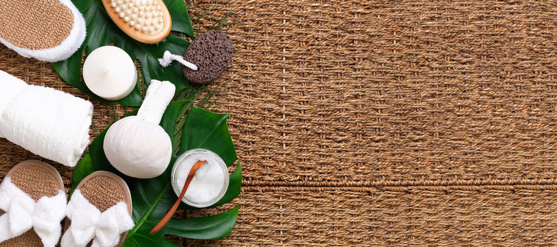 Spa tools: white towel, bamboo slippers, herbal ball, cream, wooden brush, coconut oil, monstera on rattan background. Cosmetic products for body treatment. Beauty, relax and massage concept