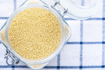 Uncooked dry couscous in a glass jar on a white checkered towel, top view, copy space.