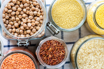 Various raw grains, flakes and legumes in glass jars, top view. Uncooked chickpeas, polenta, couscous and rice on a white checkered tablecloth.