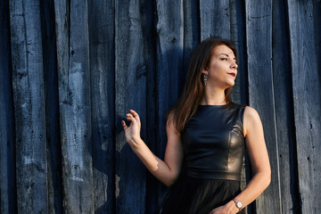 Fototapeta premium Portrait of a young slim woman brunette in a black dress near a wooden fence. Summer sunny evening. Close-up.