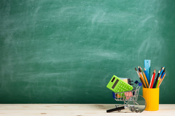 Education concept - school supplies in a shopping cart on the desk in the auditorium, blackboard...