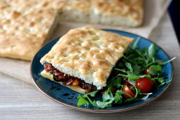 Foccacia bread sandwich with pesto, porchetta meat, eggplant and tomato sauce, served with arugula and cherry tomato salad. Homemade italian cuisine. Selective focus.
