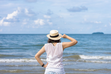 Outdoor summer portrait of Young Asian woman wearing stylish hat and clothes standing on the beach, enjoying looking view of sea with blue sky on summer vacation.