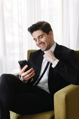 Photo of happy brunette businessman making video call on cellphone while sitting on armchair in hotel apartment