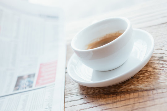 Still life image of cup of black coffee with fresh sport newspaper on the wooden table next to big coffee shop window.
