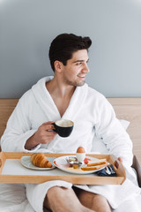 Fototapeta na wymiar Photo of unshaven caucasian man having breakfast while sitting on bed in hotel apartment