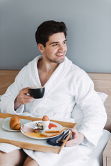 Fototapeta na wymiar Photo of happy caucasian man wearing white bathrobe having breakfast in hotel apartment
