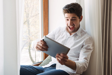 Photo of excited european man playing game on tablet computer while sitting at window in living room