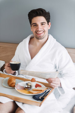 Photo of happy smiling man wearing white bathrobe having breakfast and reading newspaper in hotel apartment